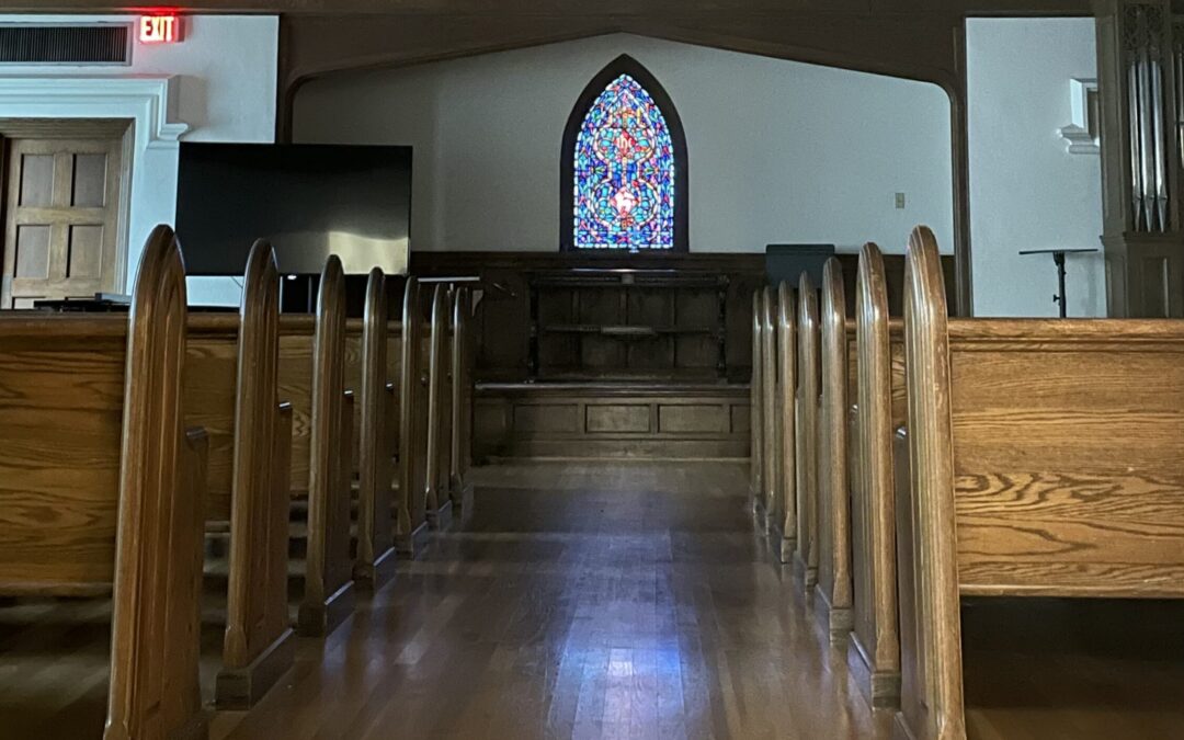 A chapel aisle lined with empty pews and looking toward a stained glass window